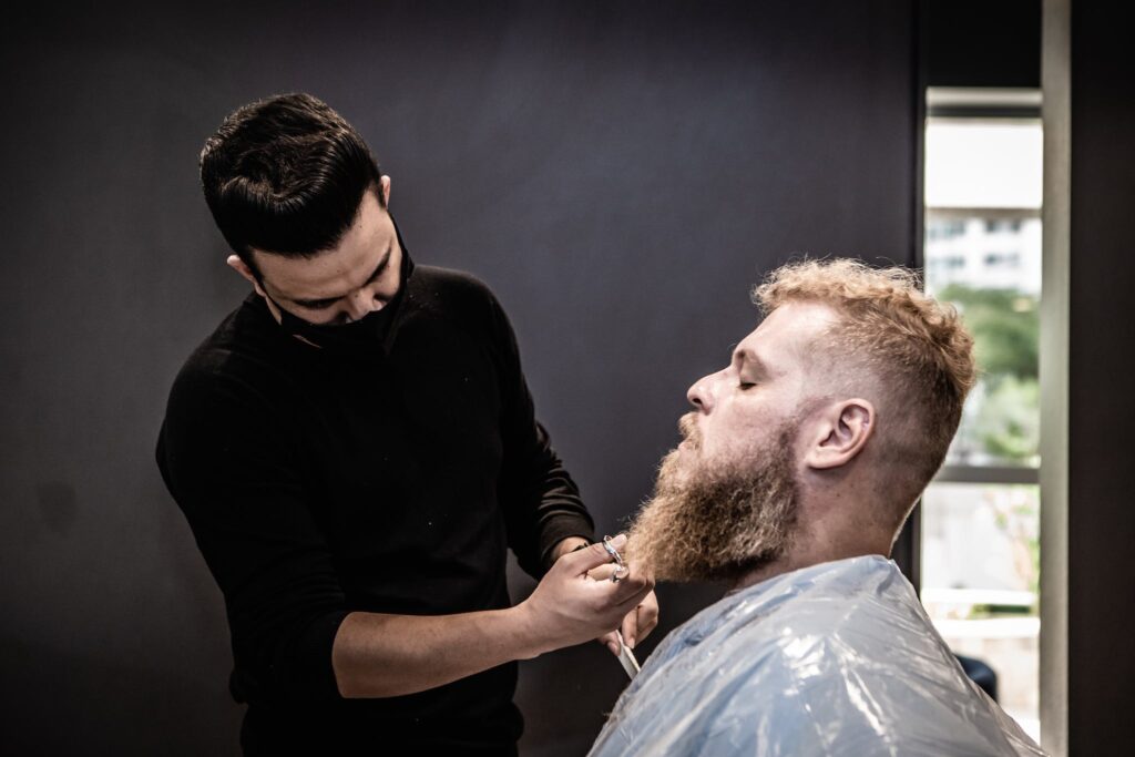 A Barber in Black Long Sleeves Trimming a Man's Beard