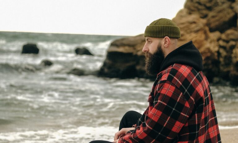 Bearded Man in Knitted Hat Sitting on Sandy Beach and Looking at Sea